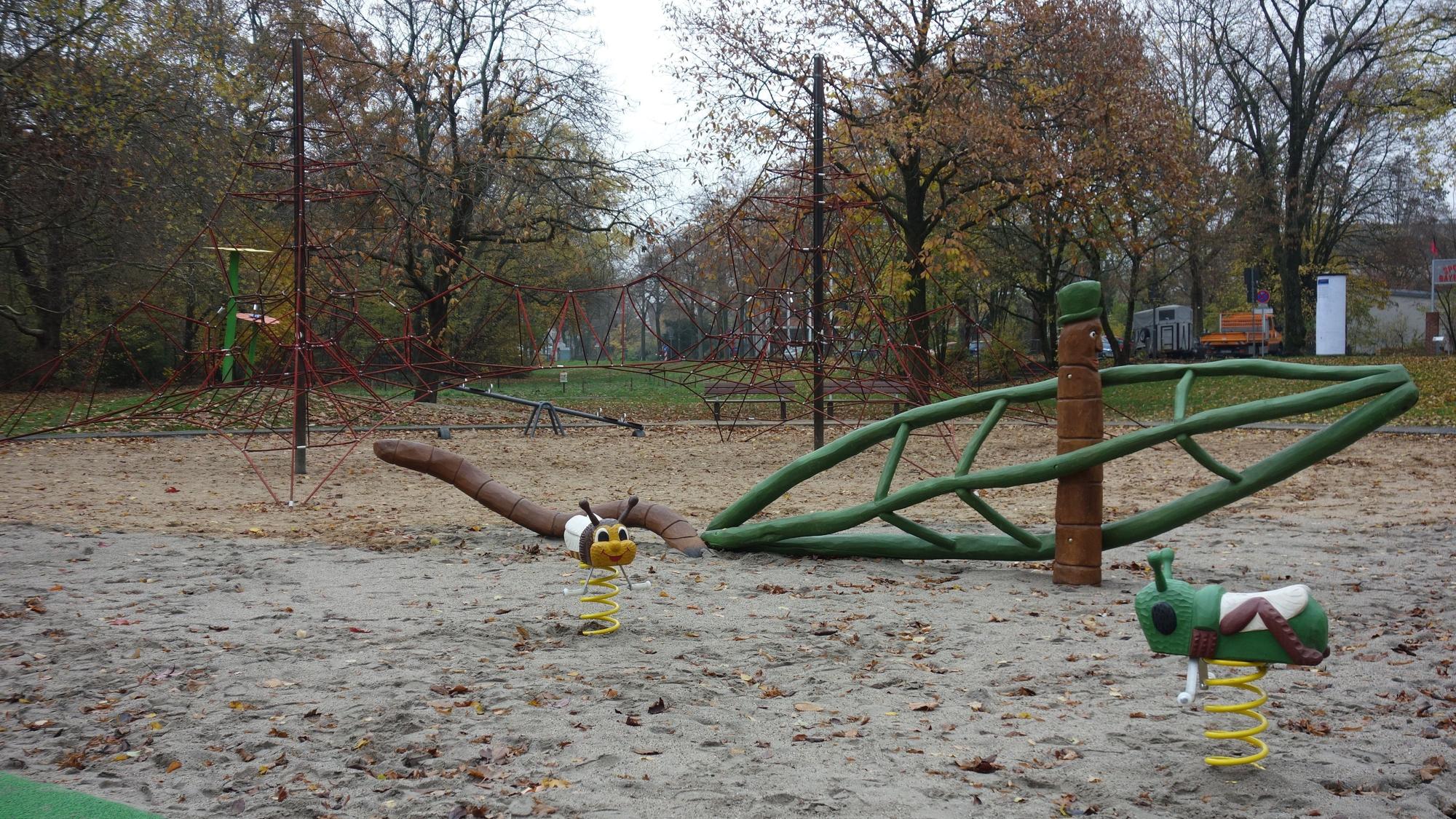 Geräte auf dem Spielplatz „Am Stadtpark“: ein rotes Seil-Klettergerüst, davor ein übergroßer Holzregenwurm sowie Federwippen in Form einer Biene und eines Grashüpfers.