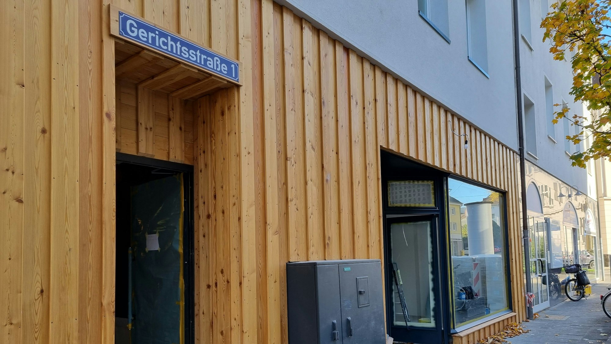 Eine holzvertäfelte Häuserfassade mit einem Schaufenster. Ein paar Meter links daneben befindet sich ein Eingang, darüber ist ein Schild mit der Aufschrift „Gerichtsstraße 1“ angebracht.