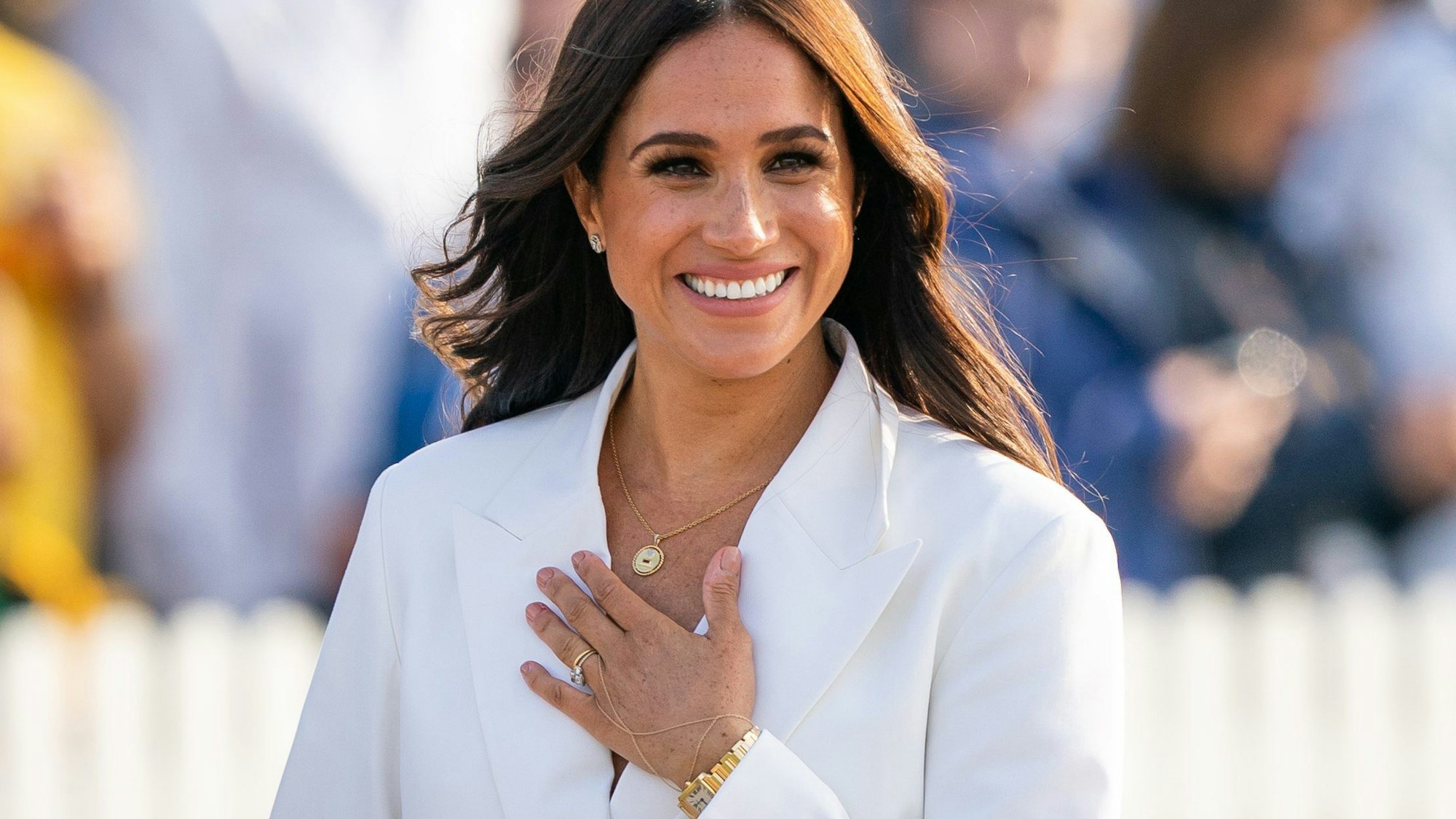 Herzogin Meghan von Sussex steht lächelnd in einem weißen Blazer bei einem Empfang in Den Haag. Sie hat die linke Hand auf den Brustkorb gelegt.