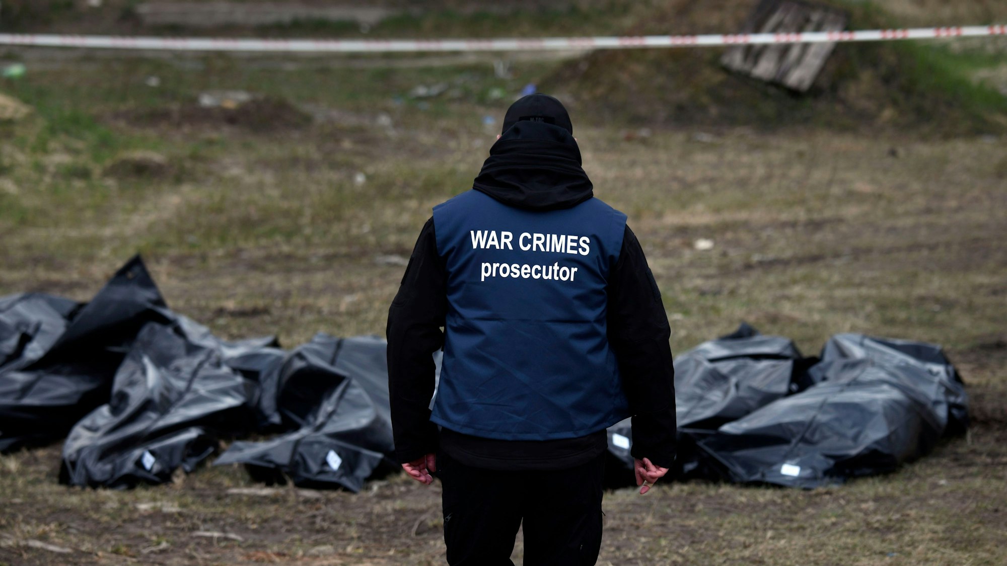 Ein Ermittler in der Ukraine trägt eine Weste mit der Aufschrift „War Crimes Prosecutor“ („Ankläger für Kriegsverbrechen“). Vor ihm liegen mehrere Leichen. (Archivbild)