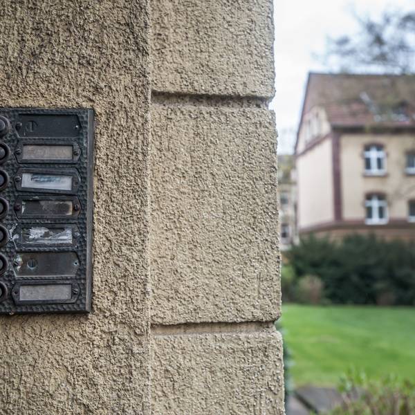 Unleserliche Klingelschilder neben einer Haustür in einem Wohngebiet. Im Hintergrund ist eine Wiese und ein weiteres Wohnhaus zu erkennen.