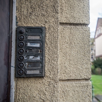 Unleserliche Klingelschilder neben einer Haustür in einem Wohngebiet. Im Hintergrund ist eine Wiese und ein weiteres Wohnhaus zu erkennen.