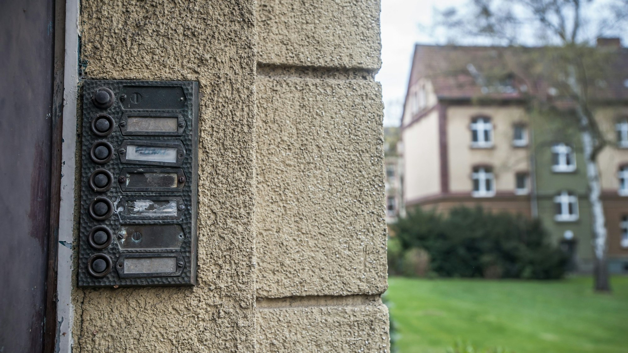 Unleserliche Klingelschilder neben einer Haustür in einem Wohngebiet. Im Hintergrund ist eine Wiese und ein weiteres Wohnhaus zu erkennen.