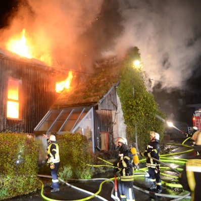 Feuerwehrleute stehen vor einem Haus mit Holzverkleidung, das in Flammen steht. Das Feuer kommt aus einem Fenster und dem Dach. In einem anderen Fenster ist deutlich zusehen, wie es im Inneren des Hauses brennt.