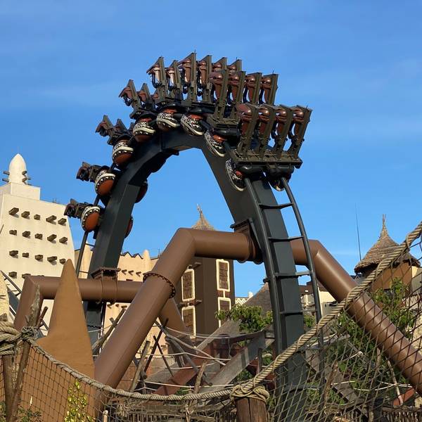 Eine Achterbahn hängt in einem Looping über Kopf. Im Hintergrund stehen Gebäude aus dem Themenreich Afrika im Phantasialand.