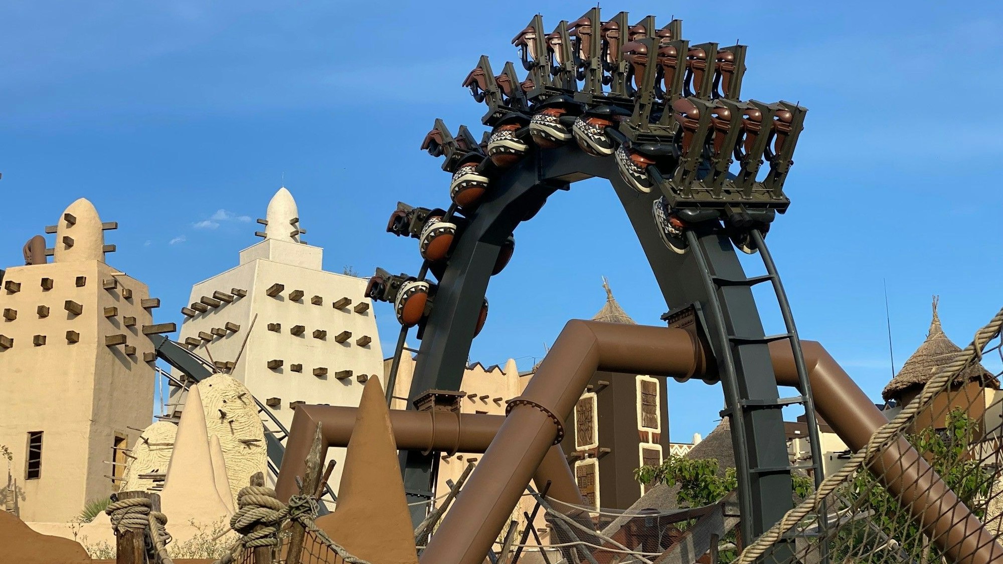 Eine Achterbahn hängt in einem Looping über Kopf. Im Hintergrund stehen Gebäude aus dem Themenreich Afrika im Phantasialand.