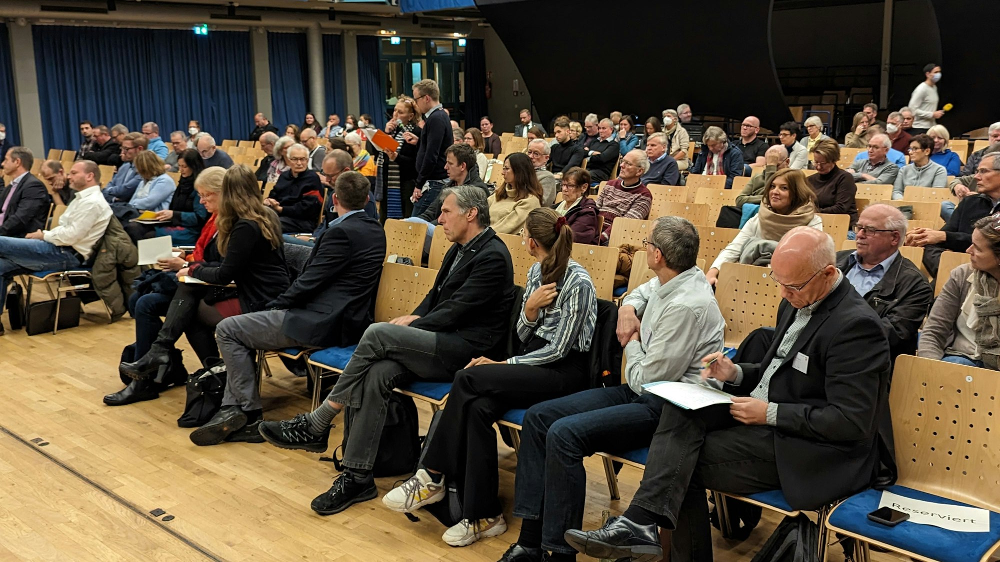 In einer Halle stehen mehrere Stuhlreihen. Viele Personen sitzen auf den Stühlen.