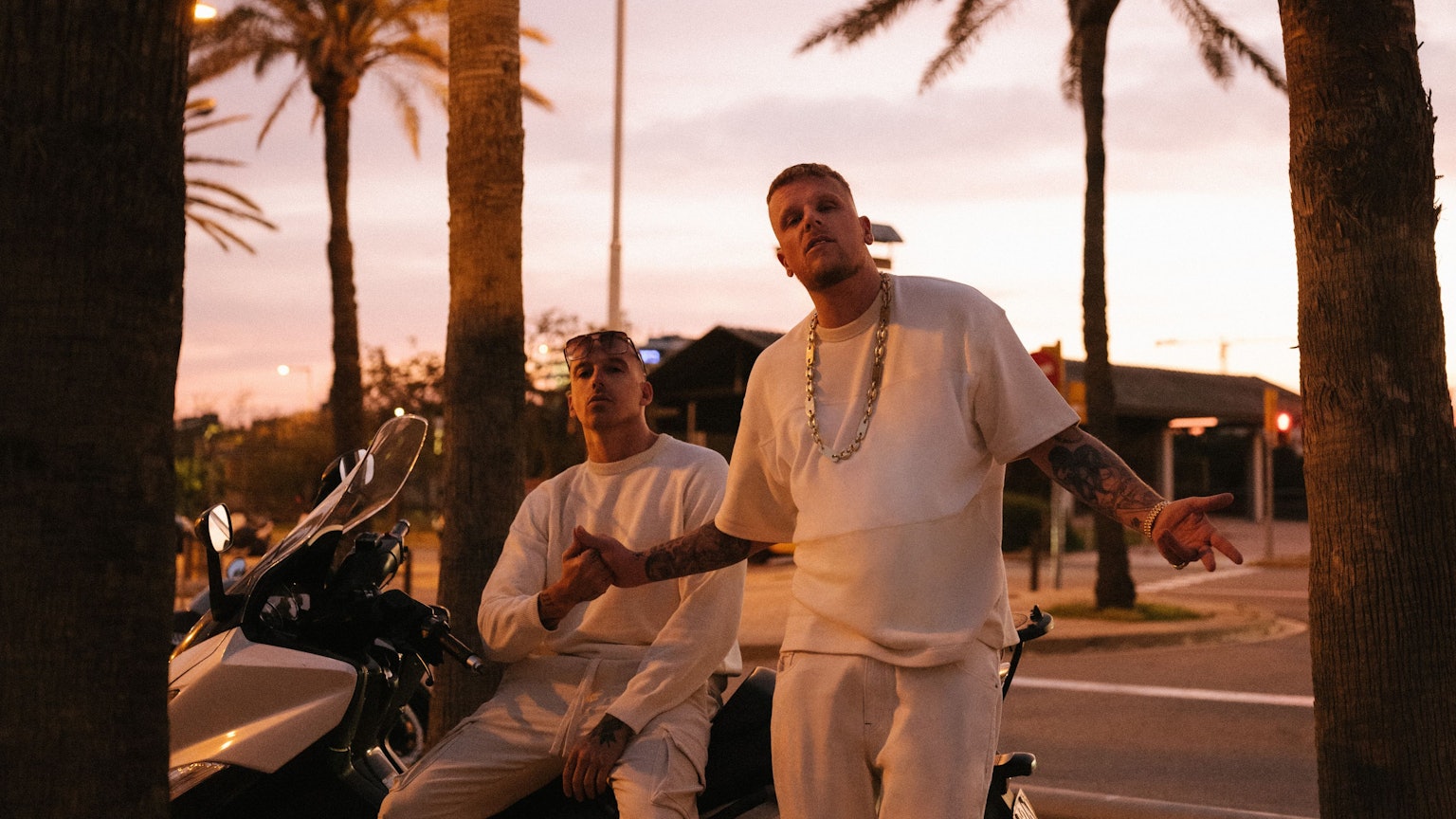 Die Rapper Bonez MC & RAF Camora in lässiger Pose unter Palmen auf Motorrädern. Sie halten sich die Hand. Sie tragen weiße Sportsachen.