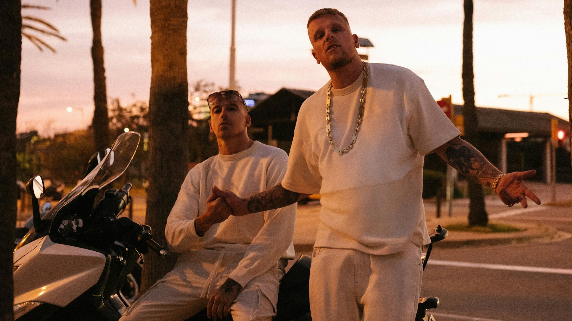 Die Rapper Bonez MC & RAF Camora in lässiger Pose unter Palmen auf Motorrädern. Sie halten sich die Hand. Sie tragen weiße Sportsachen.