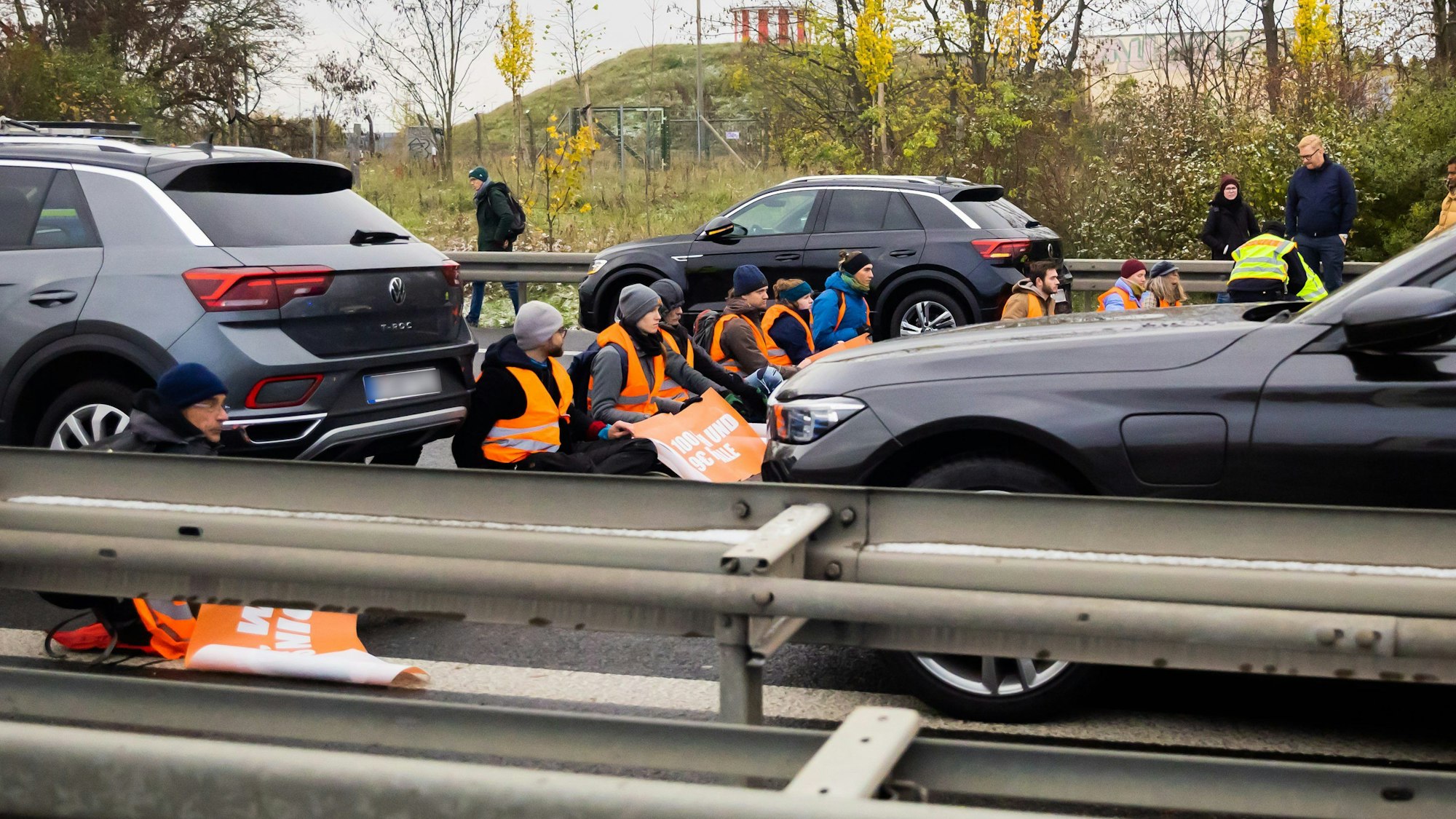 In der Bildmitte sitzen Klimaaktivisten in orangen Leuchtwesten auf der gesamten Fahrbahnbreite einer Autobahn in einer Reihe und blockieren so den Verkehr. Vor und hinter ihnen stehen Autos.