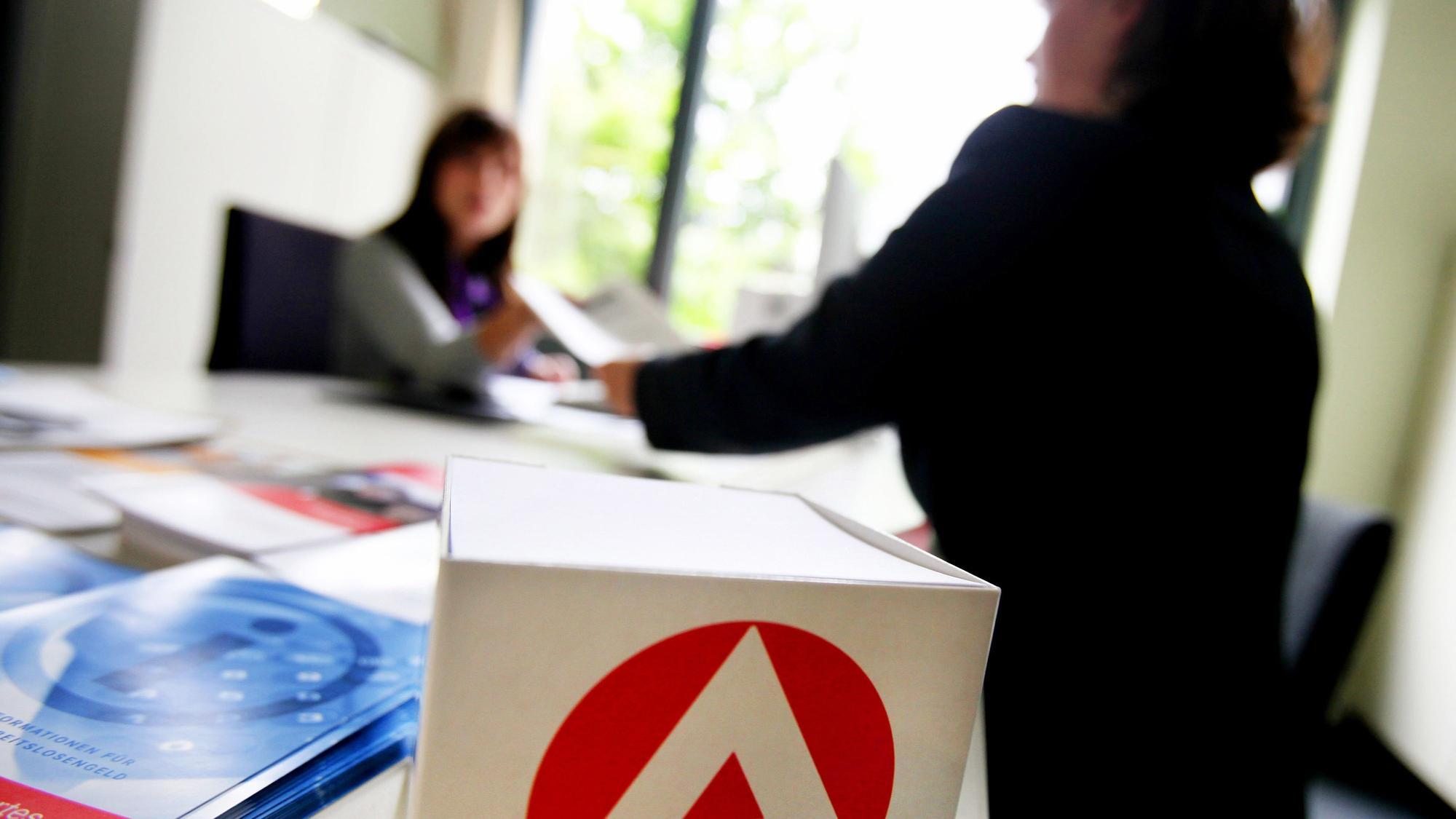 Eine Frau steht vor einem Tisch in der Arbeitsagentur, an dem eine andere Frau sitzt.
