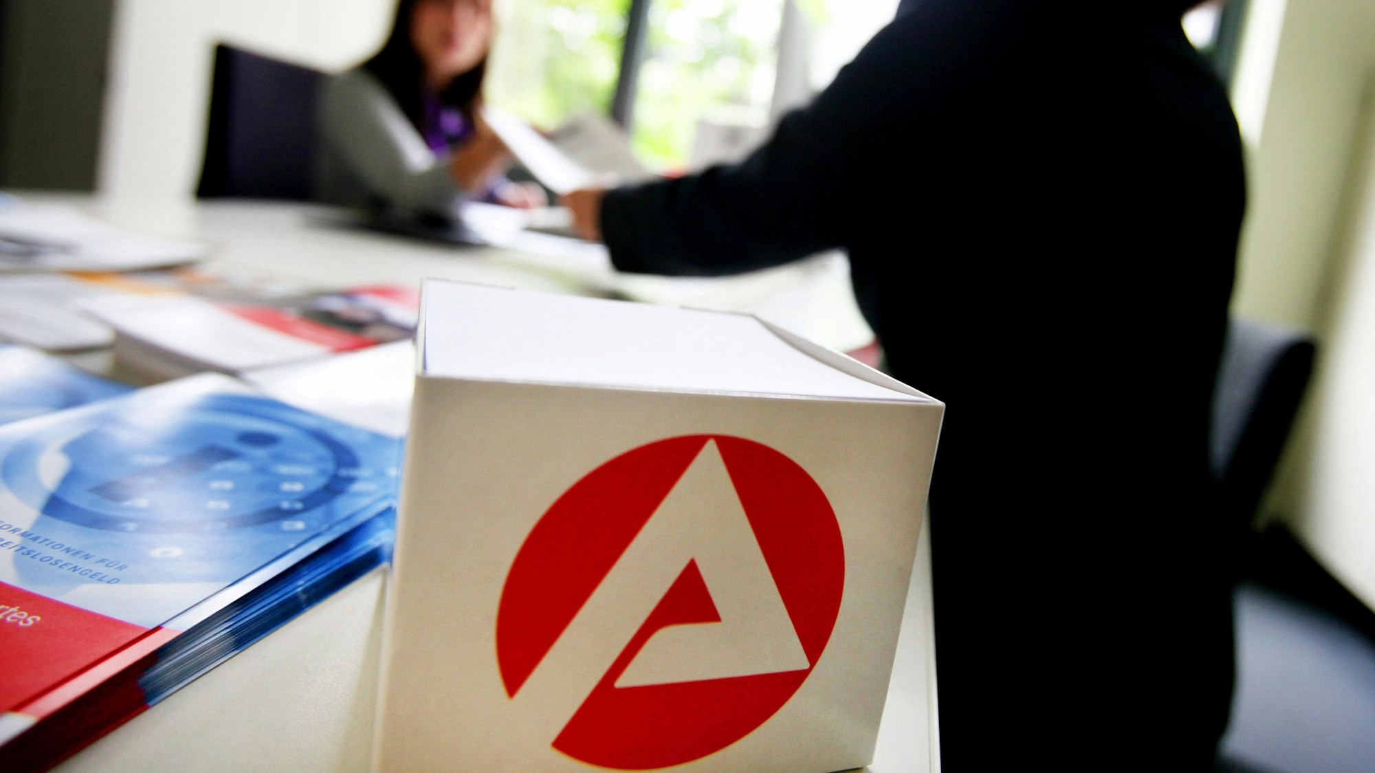 Im Vordergrund ist das Logo der Agentur für Arbeit zu sehen, im Hintergrund berät eine Mitarbeiterin der Bundesagentur für Arbeit in Düsseldorf eine Kundin.