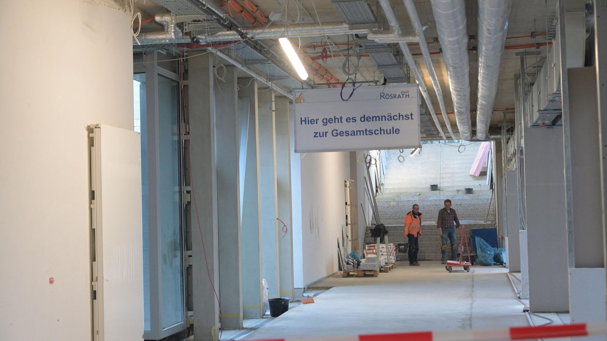 Ein noch im Bau befindlicher Flur im Rösrather Schulzentrun, an dessen Ende zwei Arbeiter stehen. An der Decke hängt ein Schild mit der Aufschrift "Hier geht es demnächst zur Gesamtschule".