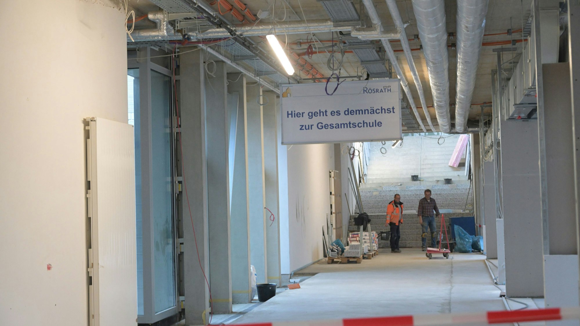 Das Bild zeigt die Baustelle, Handwerker sind noch am Arbeiten. Auf einem Schild steht „Hier geht es demnächst zur Gesamtschule“.