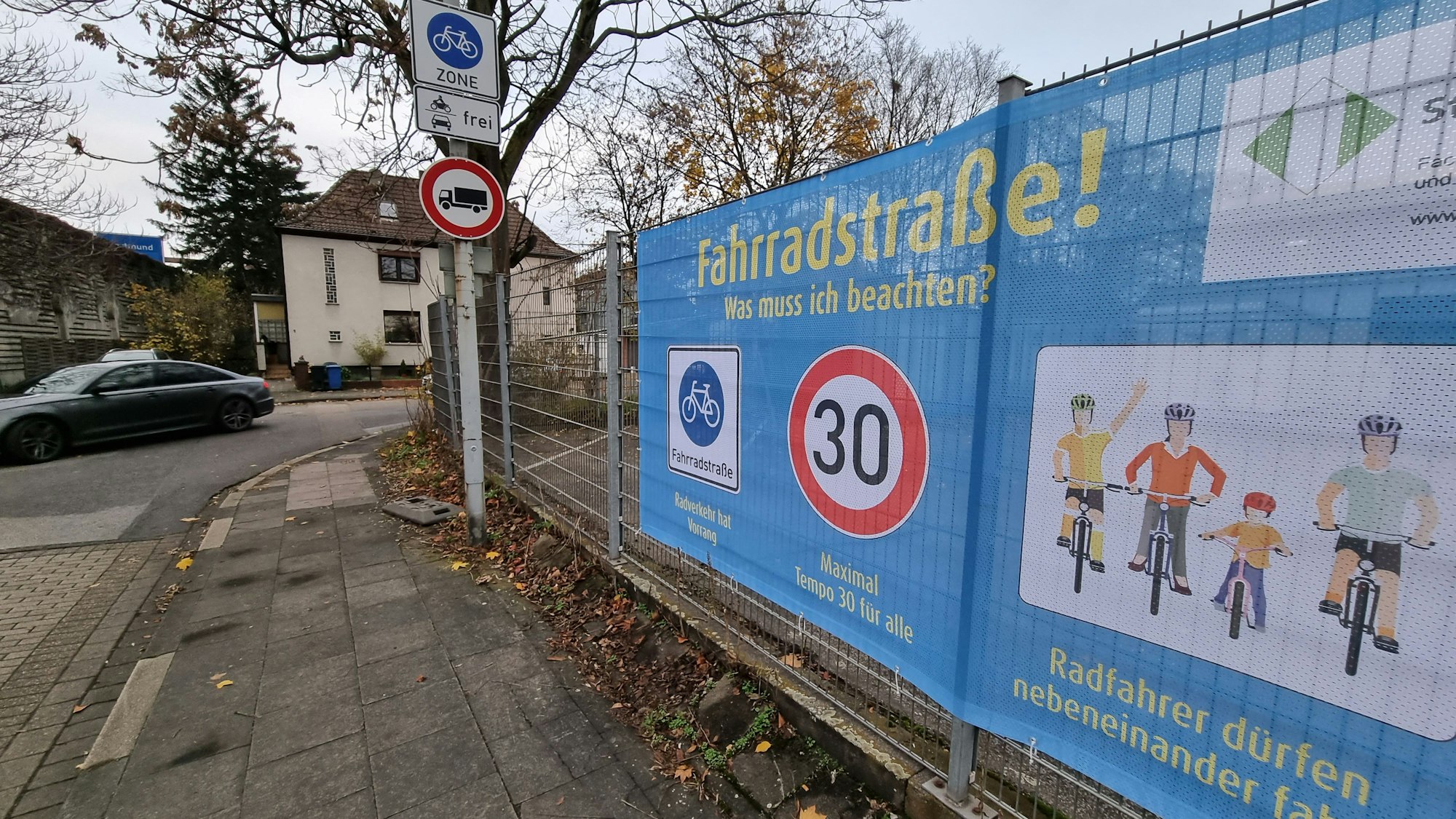 Rechts ist an einem Zaun ein großes Plakat zu sehen, auf dem die Regeln der Fahrradzone mithilfe von Piktogrammen erklärt sind. Daneben ist das Schild zu sehen, das die Fahrradzone ausweist. Ganz links biegt ein Auto um die Ecke.