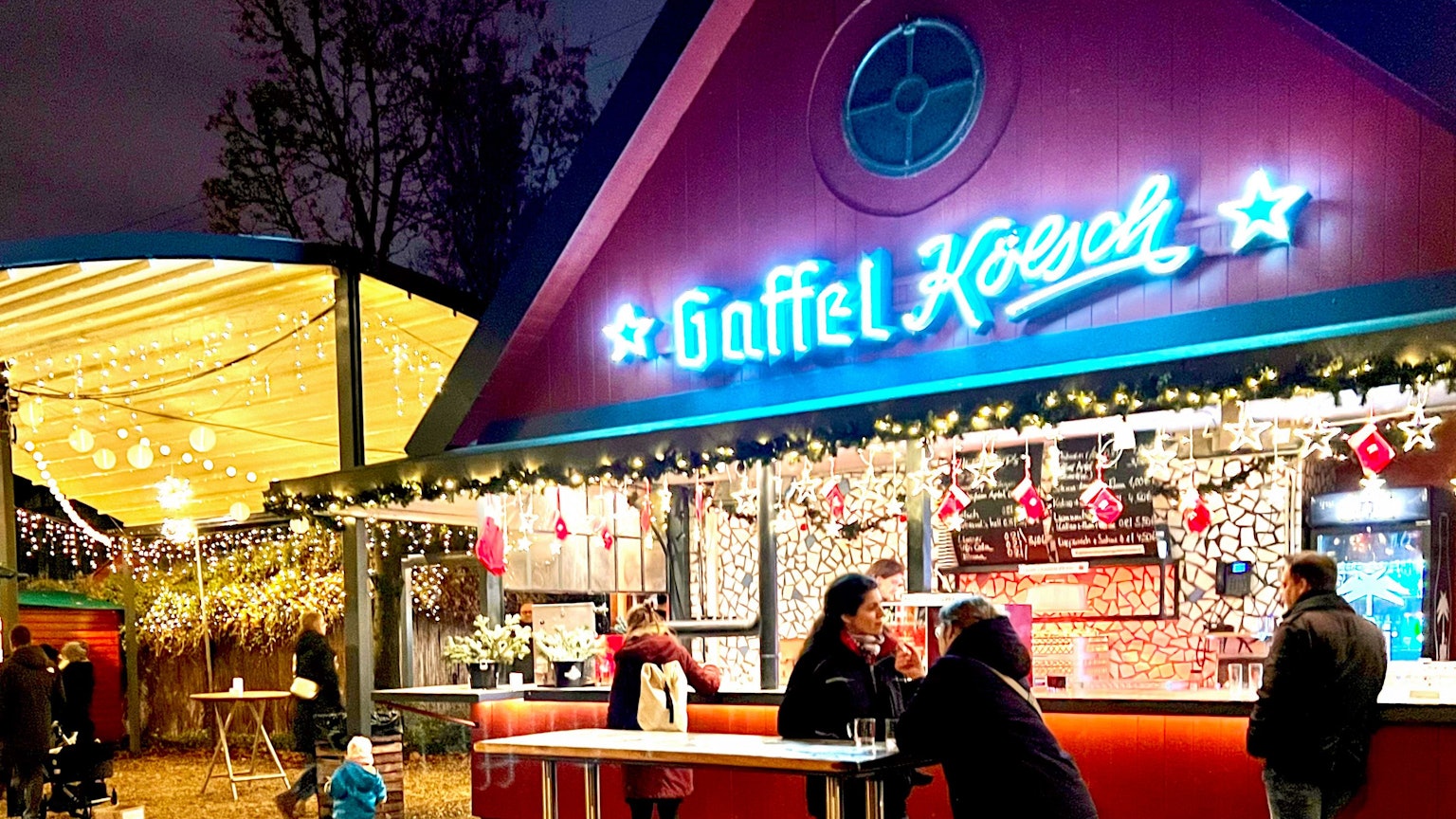 Der beleuchtete Getränke-Stand mit dem Schriftzug Gaffel Kölsch, an einem Stehtisch davor stehen zwei Frauen.