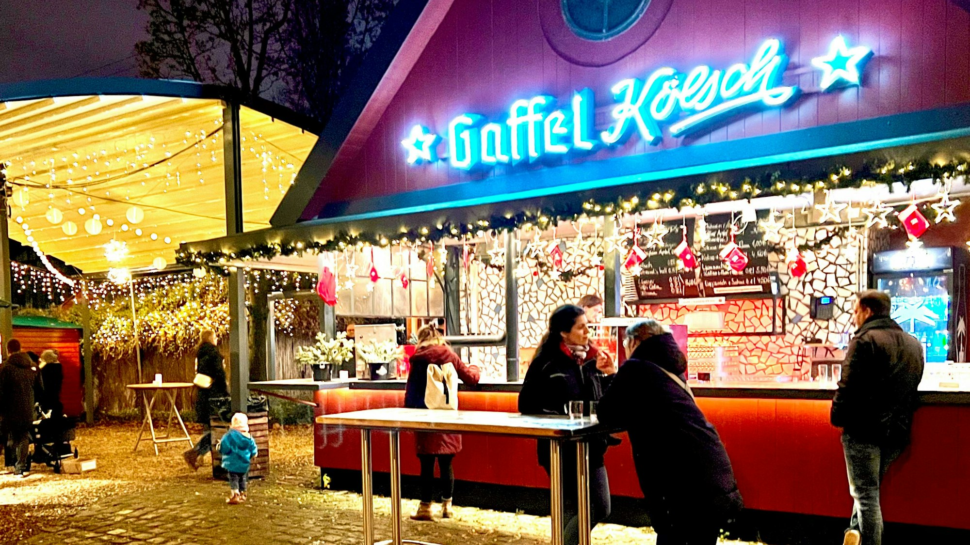 Der beleuchtete Getränke-Stand mit dem Schriftzug Gaffel Kölsch, an einem Stehtisch davor stehen zwei Frauen.