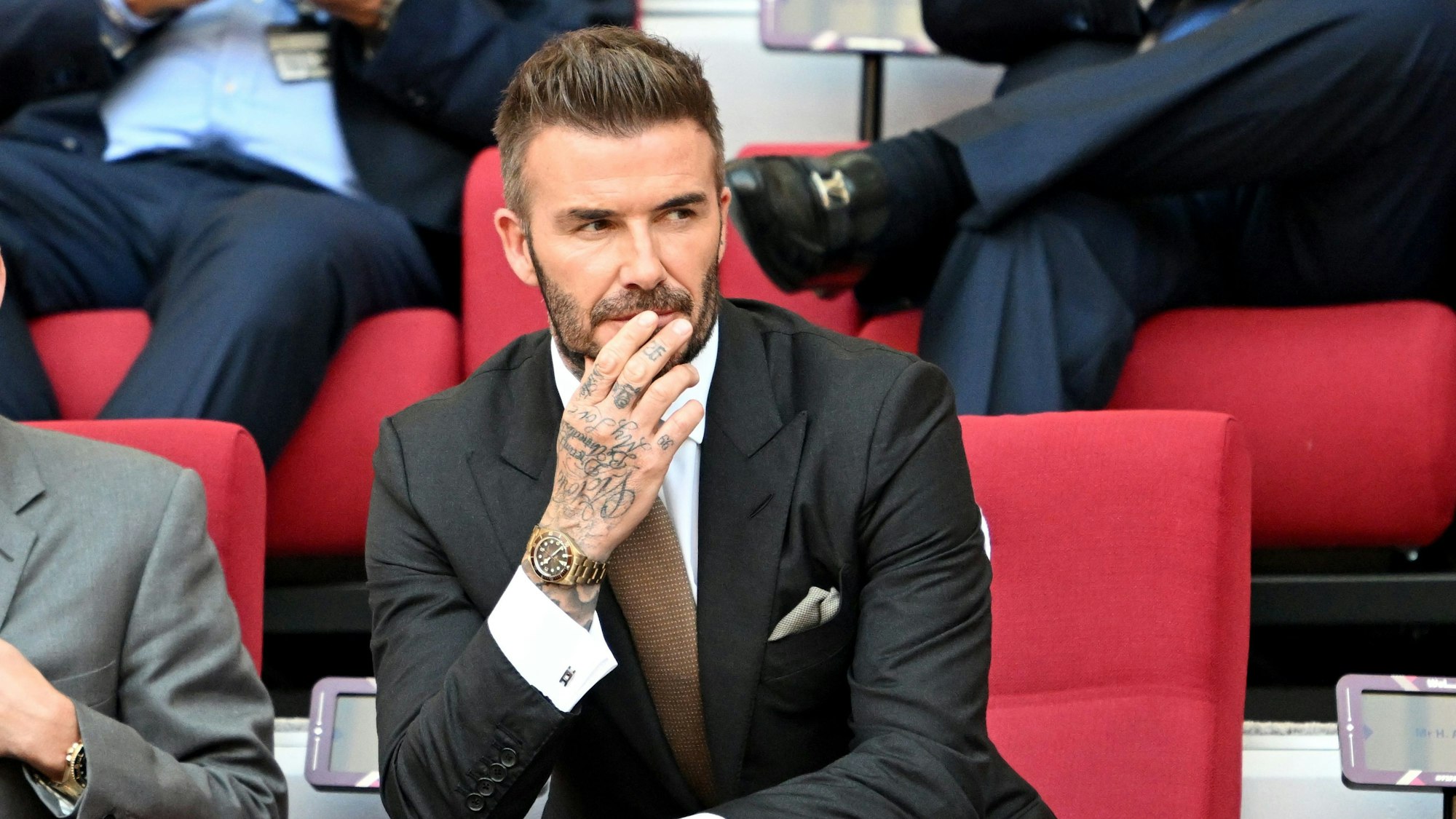 Englands David Beckham sitzt im Anzug auf der Tribüne und schaut auf das Spielfeld.