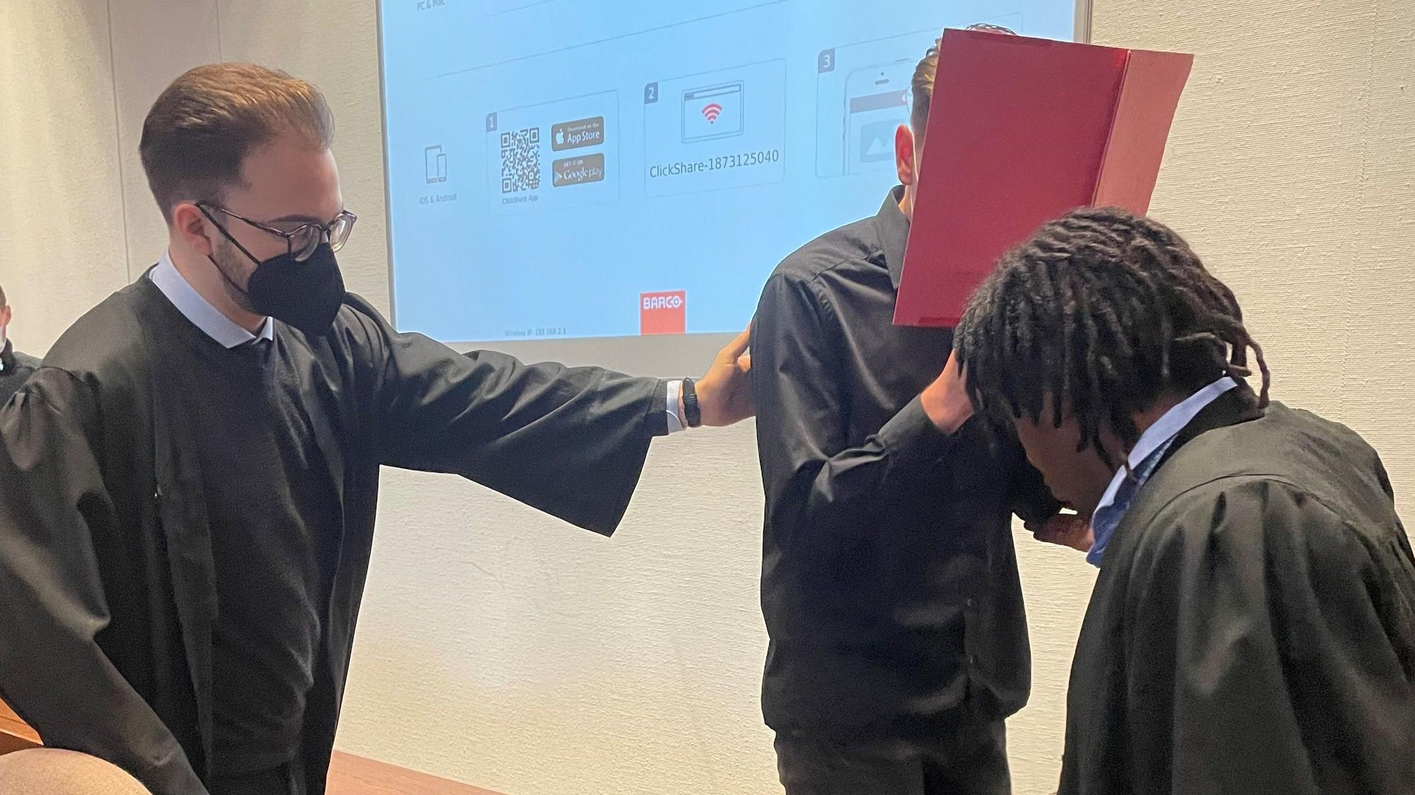 Der Angeklagte beim Prozessauftakt im Landgericht mit seinen Verteidigern Maximilian Klefenz und Dawit Stefanos. Der Angeklagte versteckt sein Gesicht hinter einem Aktenordner.
