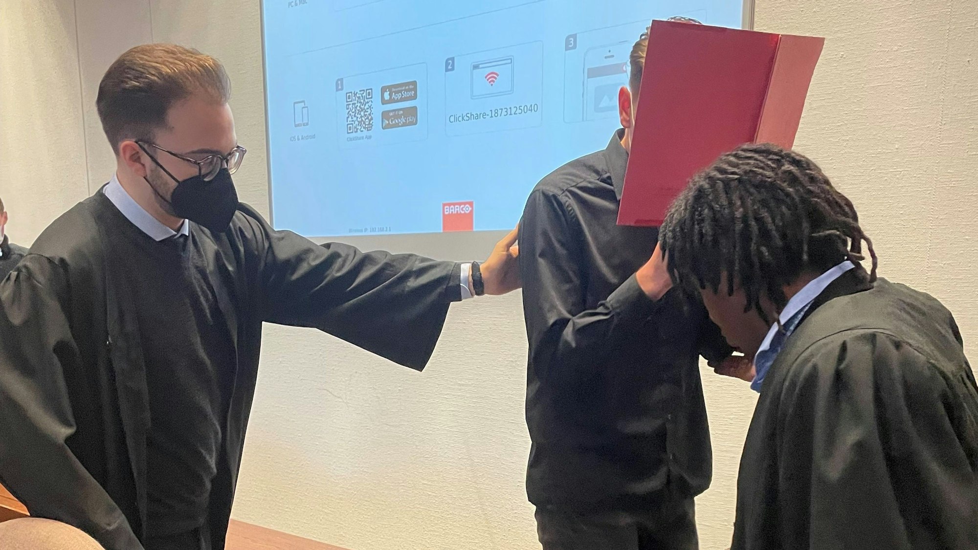 Der Angeklagte beim Prozessauftakt im Landgericht mit seinen Verteidigern Maximilian Klefenz und Dawit Stefanos. Der Angeklagte versteckt sein Gesicht hinter einem Aktenordner.