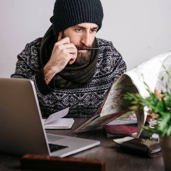 Ein Arbeitnehmer plant seinen Urlaub 2023 und hat dabei Feiertage und Brückentage fest im Blick: mit Kalender und Laptop am Schreibtisch. Er trägt Mütze, Bart und Schal und schaut konzentriert.