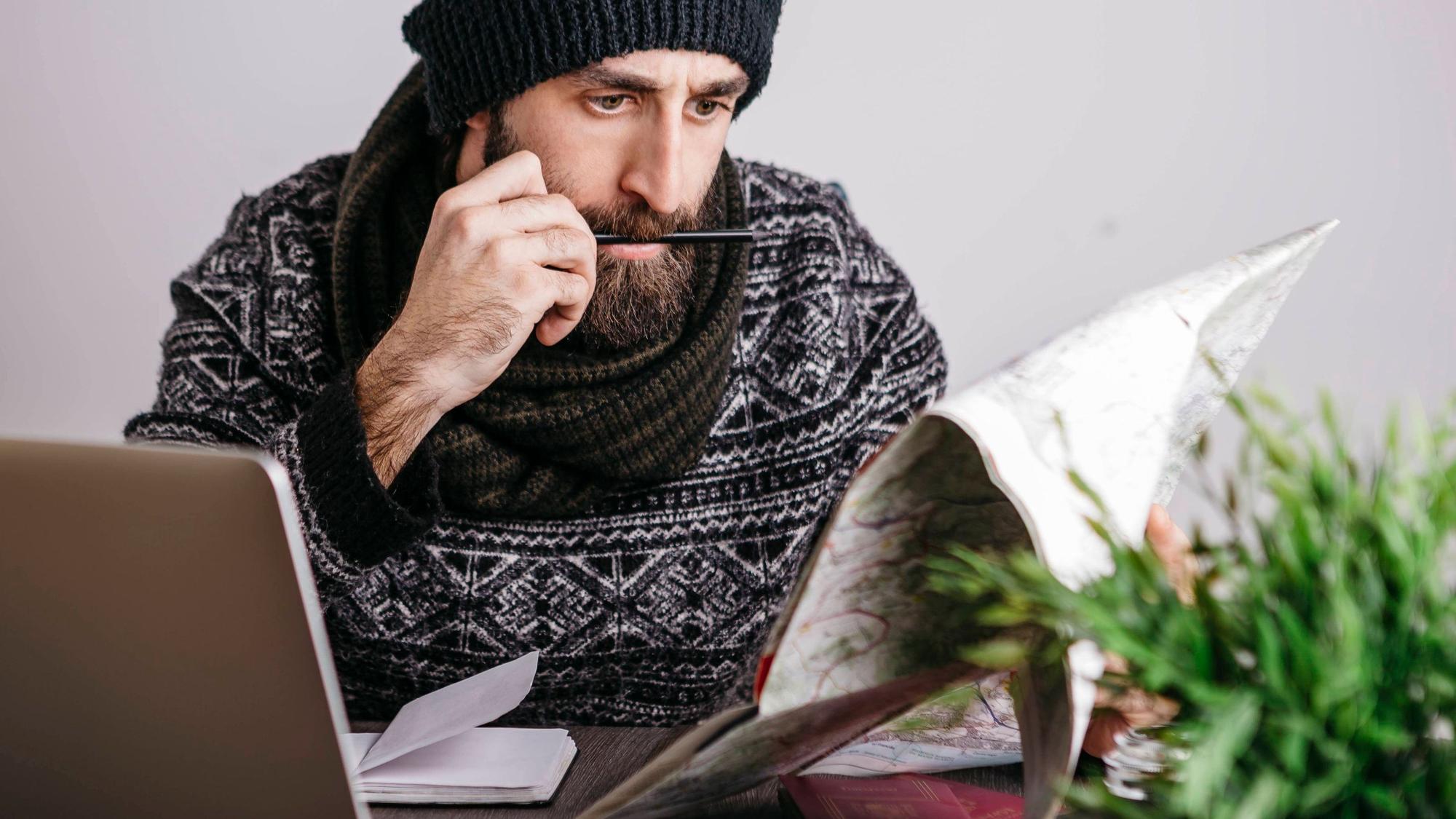 Ein Arbeitnehmer plant seinen Urlaub 2023 und hat dabei Feiertage und Brückentage fest im Blick: mit Kalender und Laptop am Schreibtisch. Er trägt Mütze, Bart und Schal und schaut konzentriert.