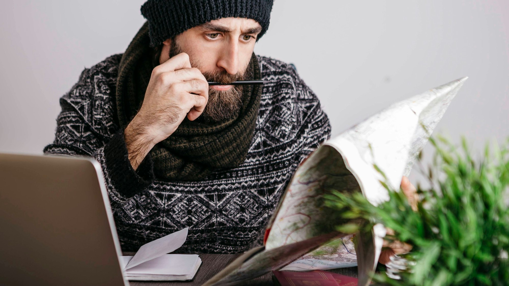 Ein Arbeitnehmer plant seinen Urlaub 2023 und hat dabei Feiertage und Brückentage fest im Blick: mit Kalender und Laptop am Schreibtisch. Er trägt Mütze, Bart und Schal und schaut konzentriert.