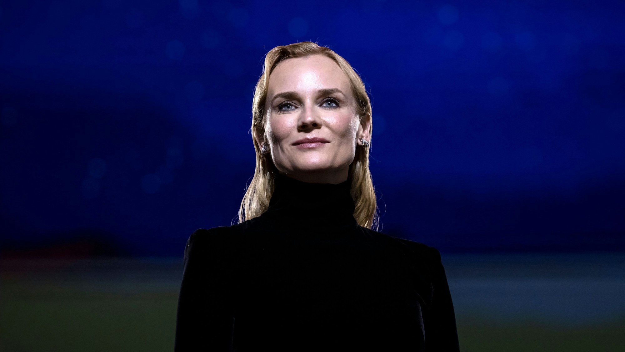 Es ist Diane Kruger vor einem blauen Hintergrund zu sehen. Sie trägt schwarze Kleidung.