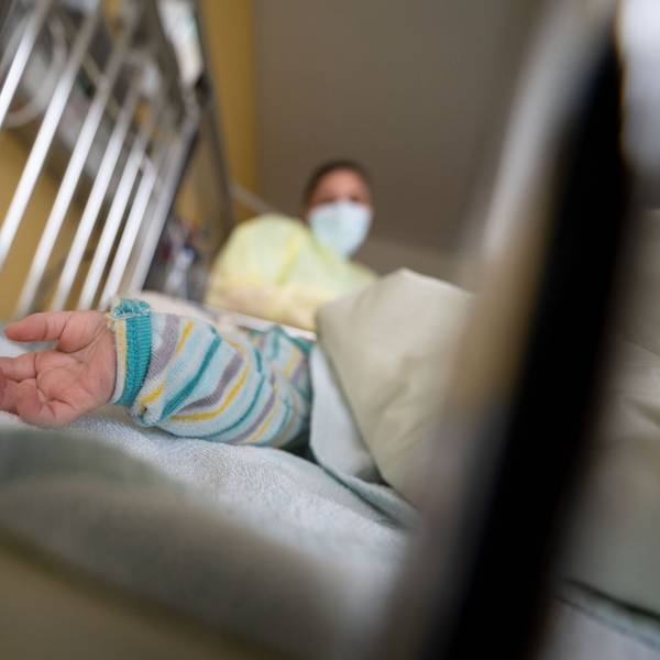 In einem Gitterbett ist eine Kinderhand zu sehen. Im Hintergrund steht eine Frau mit Mundschutz und Kittel.