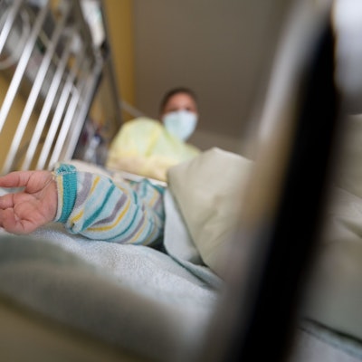In einem Gitterbett ist eine Kinderhand zu sehen. Im Hintergrund steht eine Frau mit Mundschutz und Kittel.