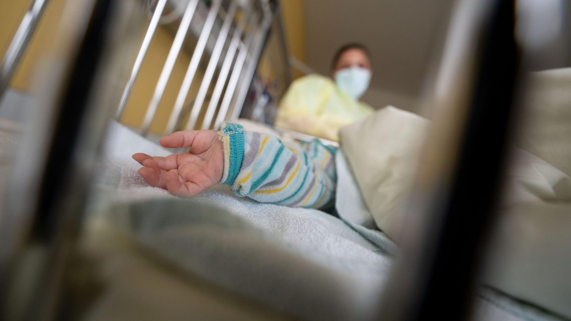 In einem Gitterbett ist eine Kinderhand zu sehen. Im Hintergrund steht eine Frau mit Mundschutz und Kittel.
