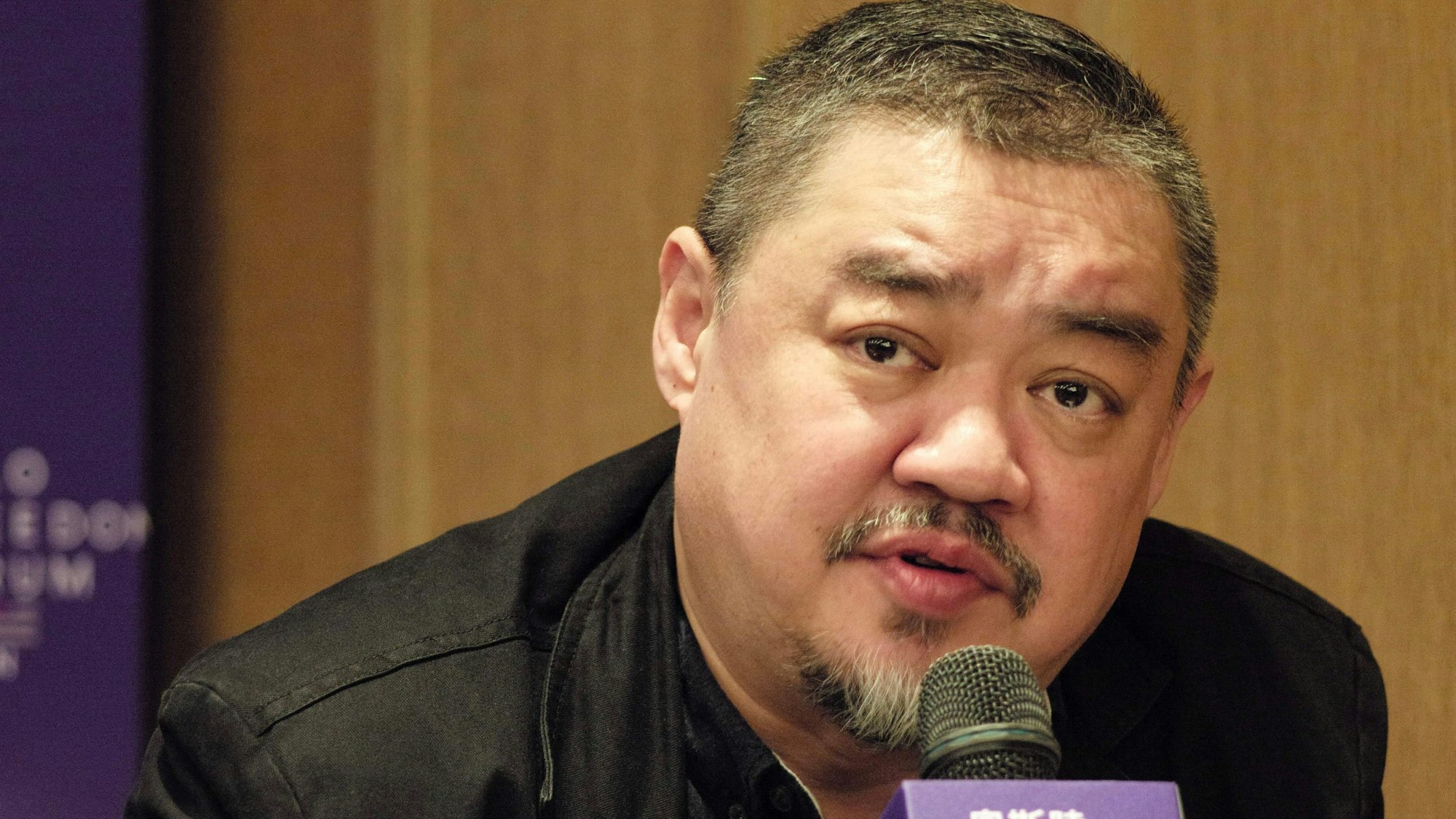 Es ist Wu'er Kaixi zu sehen, der vor einem Mikrofon sitzt.