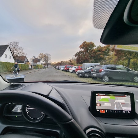 Blick über das Lenkrad eines Autos nach draußen. Zu sehen sind eine Straße, rechts parken Autos, links fährt eine Frau auf einem Fahrrad.