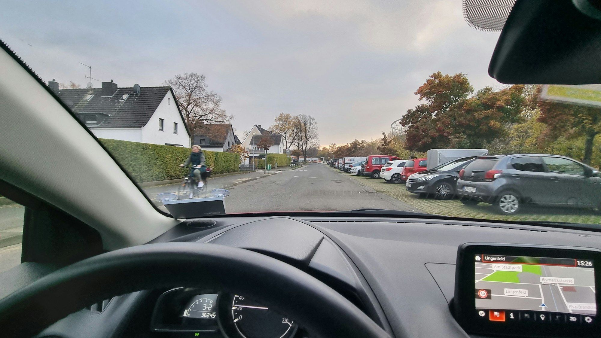 Blick über das Lenkrad eines Autos nach draußen. Zu sehen sind eine Straße, rechts parken Autos, links fährt eine Frau auf einem Fahrrad.
