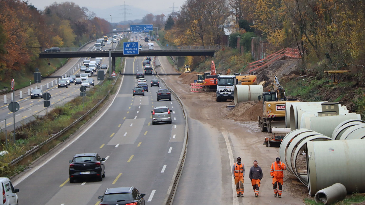 Auf einer Autobahn fahren Fahrzeuge auf drei verengten Fahrstreifen. Am rechten Fahrbahnrand laufen drei Baustellenarbeiter mit Warnkleidung über den abgesperrten Standstreifen. Im Hintergrund stehen Bagger.