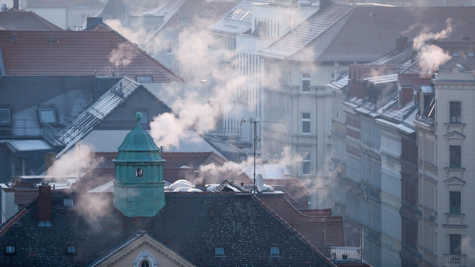 Die Dächer einer Siedlung im Winter, aus den Schornsteinen strömt Gas.