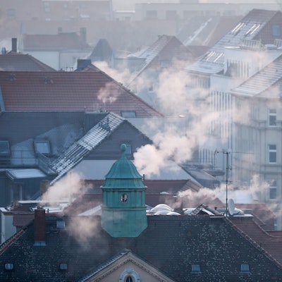 Die Dächer einer Siedlung im Winter, aus den Schornsteinen strömt Gas.