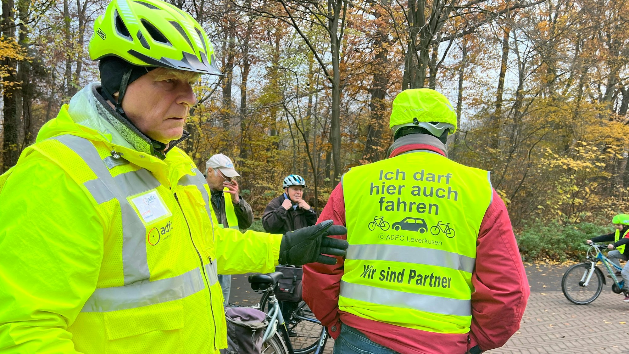 Links steht Kurt Krefft in signalgelber Jacke und mit signalgelbem Helm, mit dem Rücken zur Kamera steht ein Mann in Warnweste. Darauf steht geschrieben: „Ich darf hier auch fahren. Wir sind Partner.“