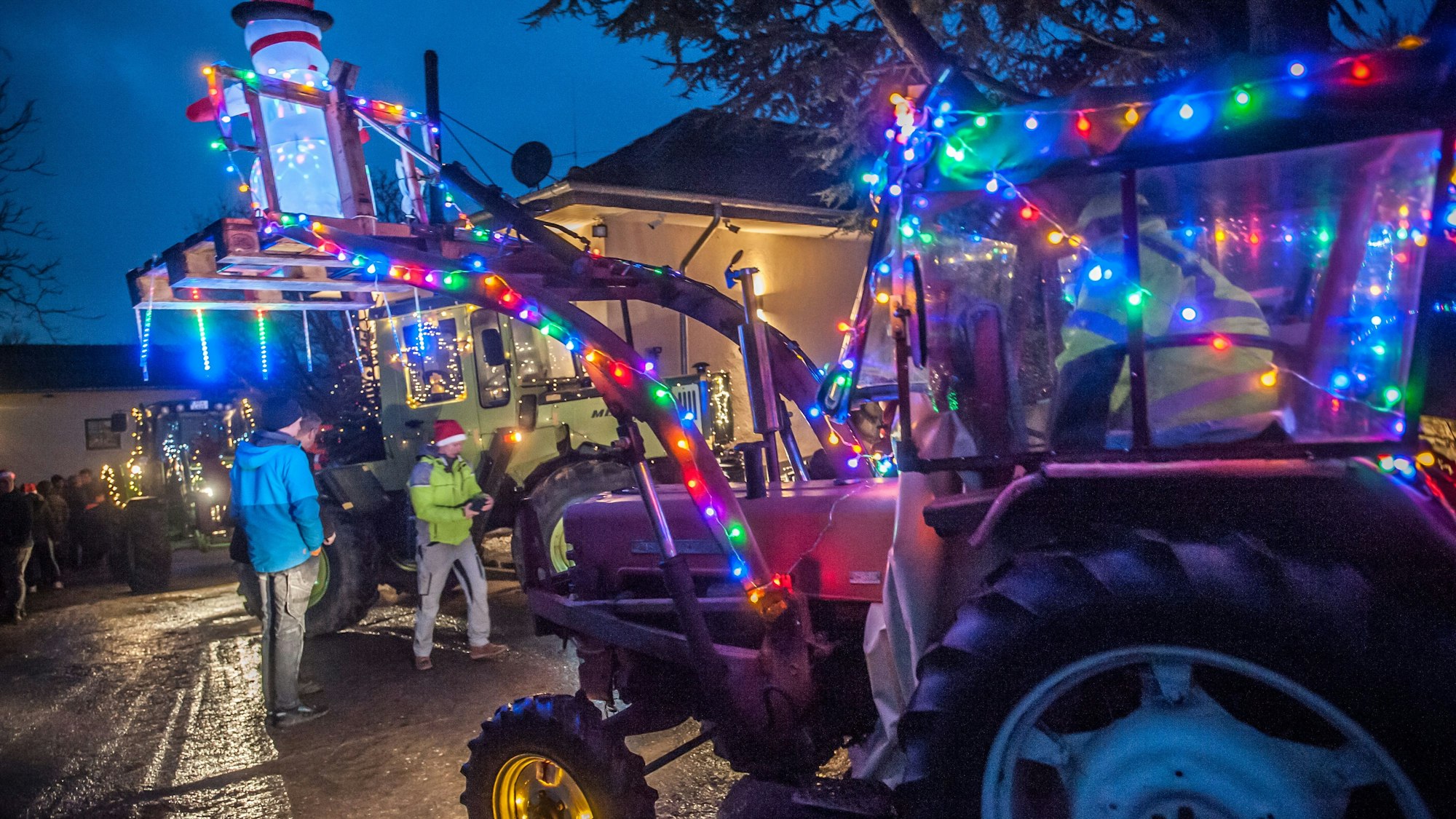 Zwei mit bunten Lichterketten und einem beleuchteten Schneemann dekorierte Traktoren fahren auf der Atzlenbacher Straße an Zuschauern vorbei.