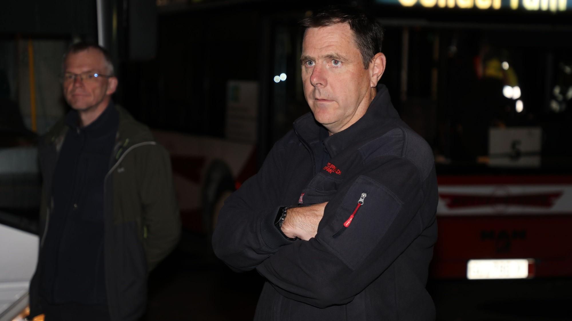 Feuerwehrchef Jörg Köhler steht im Dunkeln mit verschränkten Armen vor einem Bus, dessen Anzeige „Sonderfahrt“ anzeigt. Im Hintergrund steht Bürgermeister Frank Stein.