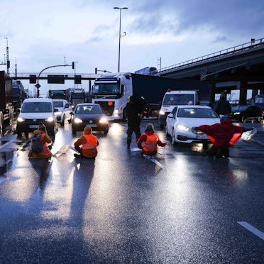 Klimaaktivisten sitzen mit dem Rücken zur Kamera auf einer mehrspurigen Kreuzung. Vor ihnen stehen Autos und LKW.