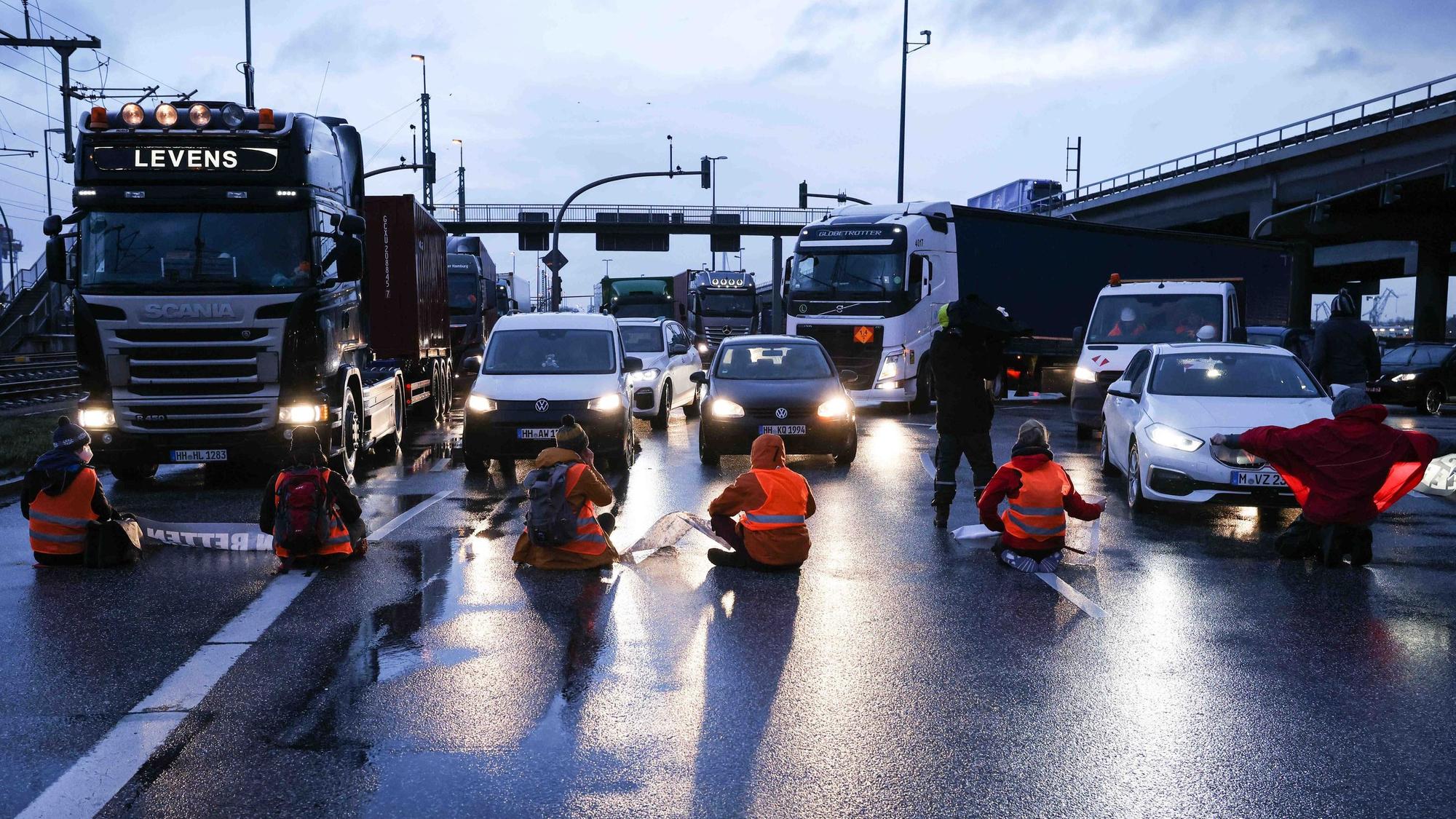 Klimaaktivisten sitzen mit dem Rücken zur Kamera auf einer mehrspurigen Kreuzung. Vor ihnen stehen Autos und LKW.