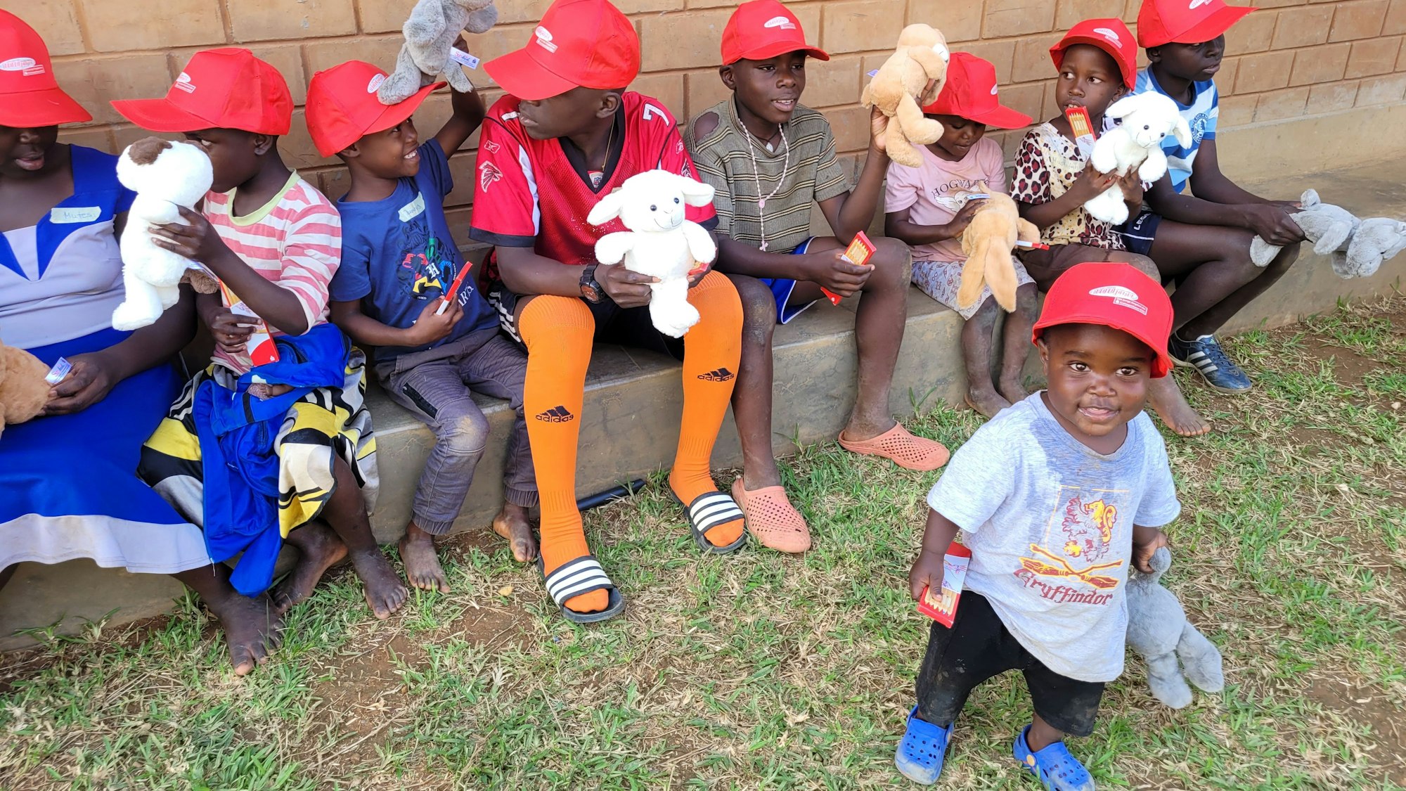 Kinder im unterschiedlichsten Alter sitzen auf einer Mauer. Alle haben ein Kuscheltier im Arm und tragen eine rotze Kappe.