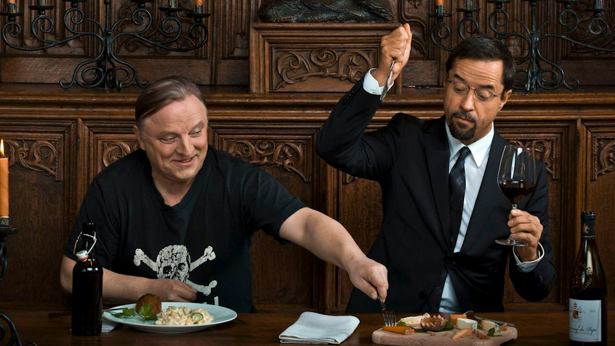 Der Pathologe Professor Karl-Friedrich Boerne (Jan Josef Liefers, rechts) sitzt Hauptkommissar Frank Thiel (Axel Prahl, links) an einem Tisch. Thiel versucht sich vom Teller Boernes etwas Essen zu nehmen. Boerne hebt das Messer in der einen Hand und in der anderen ein Rotweinglas.