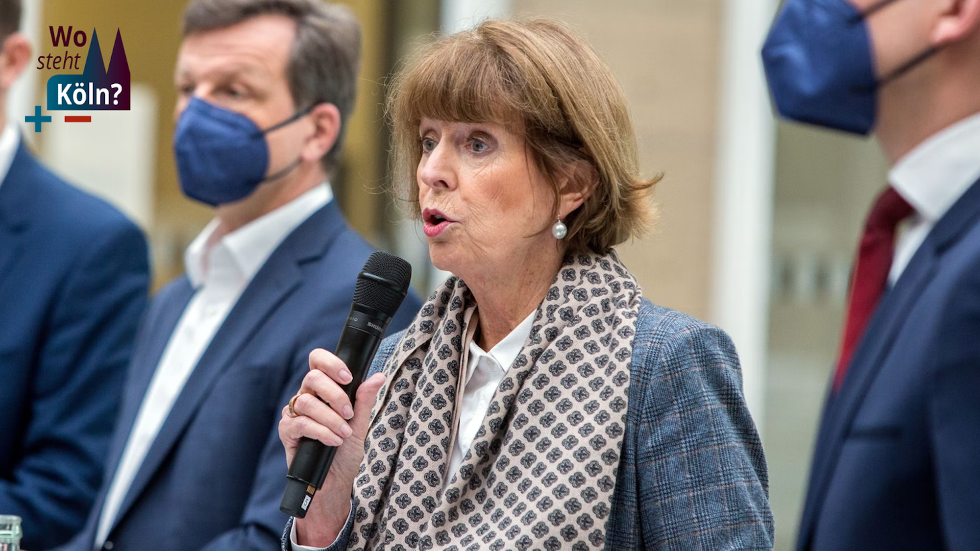 In der Bildmitte befindet sich OB Henriette Reker; sie spricht mit Mikrofon in der Hand. Rechts neben ihr steht ihr Sprecher, Alexander Vogel, links von ihr zwei Dezernenten.