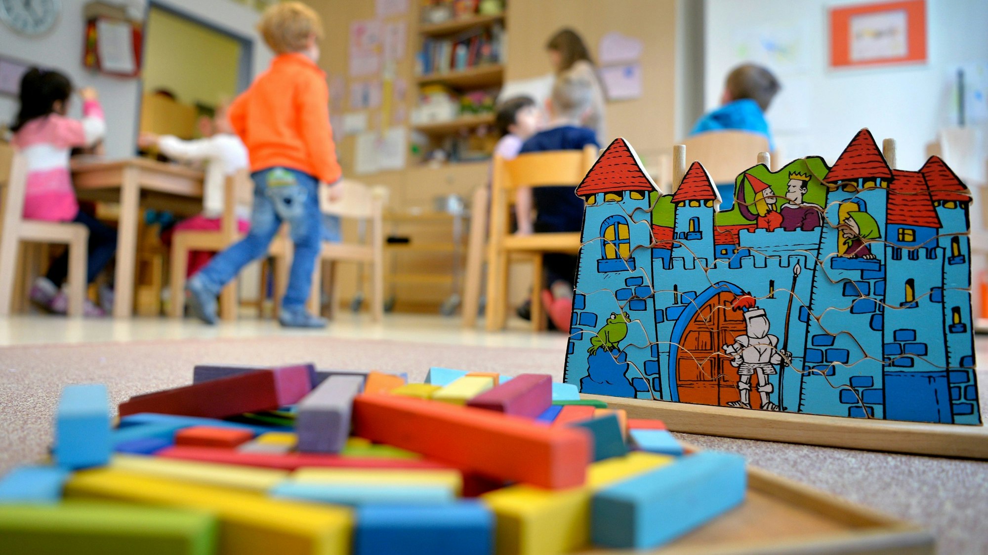 Spielzeug liegt in einer Kindertagesstätte auf dem Boden. Im Hintergrund sind Kinder zu erkennen.