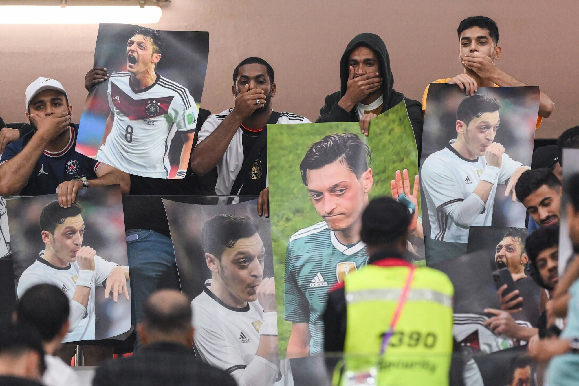 Zuschauer aus Katar halten sich den Mund zu und halten Plakate auf denen Mesut Oezil zu sehen ist in die Höhe.