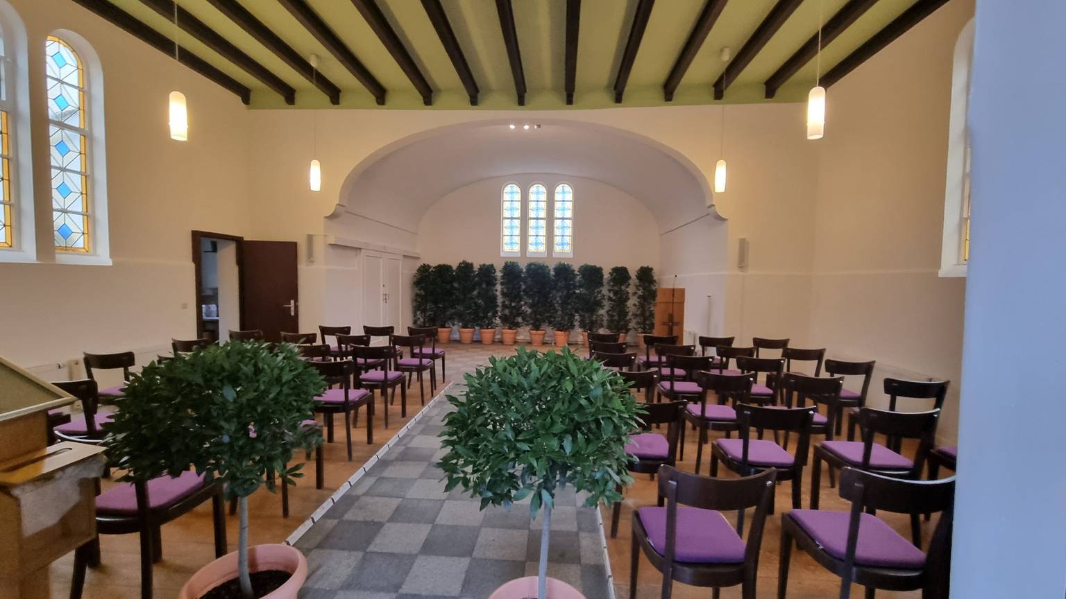 Blick in die weiß gestrichene Trauerhalle des Friedhofs in Witzhelden mit Pflanzenschmuck und leeren Stuhlreihen auf dem seitlichen Holzboden.