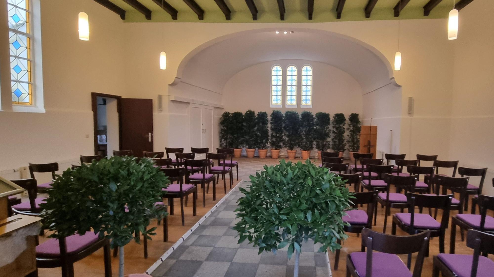 Blick in die weiß gestrichene Trauerhalle des Friedhofs in Witzhelden mit Pflanzenschmuck und leeren Stuhlreihen auf dem seitlichen Holzboden.
