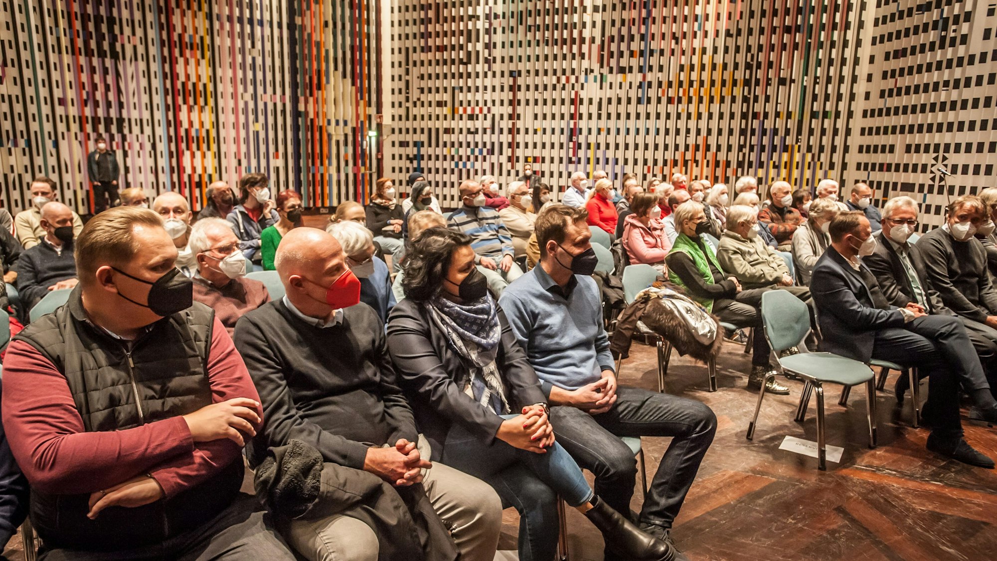 Rund 70 Personen sitzen im Agam-Saal auf Stühlen. Sie alle tragen eine FFP2-Maske.